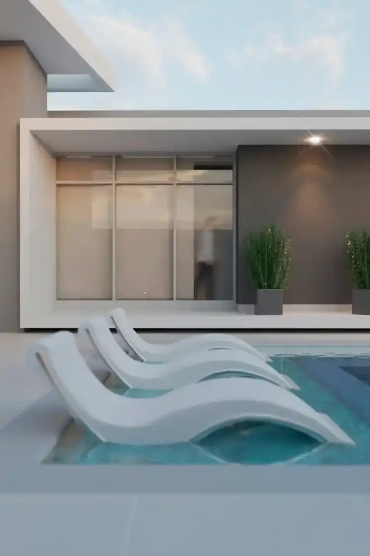 Vista en primer plano de una piscina moderna con tres asoleadoras blancas sumergidas en una playa húmeda. Al fondo, la fachada de una casa de lujo en Salgar, Atlántico, diseñada por Edson Sánchez Arquitecto, destacando amplios ventanales, estructura blanca enmarcada y muros grises con macetas iluminadas, reflejando el estilo de vida exclusivo del Caribe