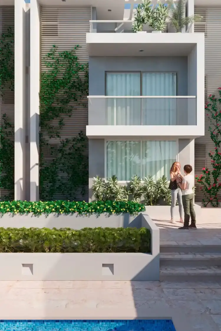 Vista vertical de la fachada de un edificio residencial moderno y sostenible en Barranquilla, diseñado por Edson Sánchez Arquitecto. La imagen muestra líneas limpias de concreto blanco, balcones con barandillas de vidrio, un jardín en la azotea, vegetación trepadora en pantallas de madera, una piscina en primer plano con mosaicos azules y dos personas conversando en la terraza pavimentada. El diseño integra naturaleza y modernidad para maximizar la calidad de vida en el Caribe